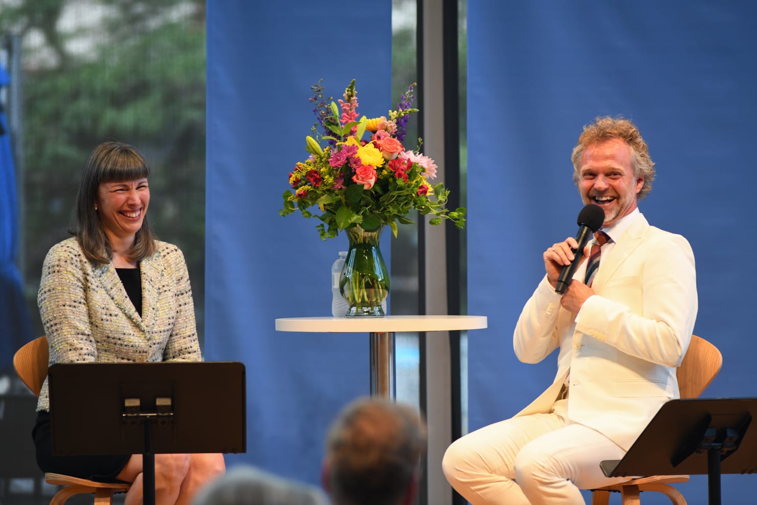 Michelle Miller Burns and Music Director Thomas Søndergård seated next to each other on a small stage, both laughing and holding microphones.